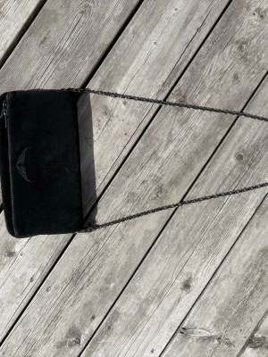 Svart Zadig Voltaire väska 💕 - Stilren svart kuvertväska i mockaliknande material med en diskret broderad detalj framtill från märket Zadig Voltaire. Väskan har en lång kedjerem i metall som gör den enkel att bära crossbody eller på axeln. Perfekt för dig som gillar minimalistisk och edgy stil. 💕