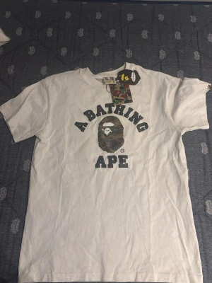 Vit A Bathing Ape t-shirt - Säljer en vit t-shirt från A Bathing Ape med det klassiska BAPE-logotrycket och kamouflagemönstrad apa på bröstet. T-shirten har rund hals och korta ärmar. Perfekt för dig som gillar streetwear och vill sticka ut med en ikonisk design.