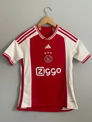 Ajax fotbollströja röd/vit Adidas -  Ajax hem tröja med sponsorer Ziggo på magen. Köpt på Adidas officiella butik och har blivit för liten för mig. Storlek 11-12 eller 152. Perfekt för dig som gillar streetwair eller samlar på snygga fotbollströjor.