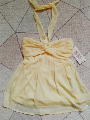 Ljusgul bandeau topp med knyt - Supersöt ljusgul bandeau topp från Nelly med knyt runt nacken och twistad detalj framtill. Toppen är axelbandslös och har ett luftigt, skirt tyg som ger en romantisk känsla. Perfekt för varma dagar och somriga outfits. Stl Xs. Ny med prislapp, säljer då den inte kommit till användning!