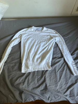 Vit långärmad tröja från Zara - Snygg vit långärmad tröja från Zara i tunnare material. Tröjan har rund halsringning och dekorativa knappar vid ärmslutet på ena ärmen. Perfekt att styla med jeans eller kjol för en clean look.