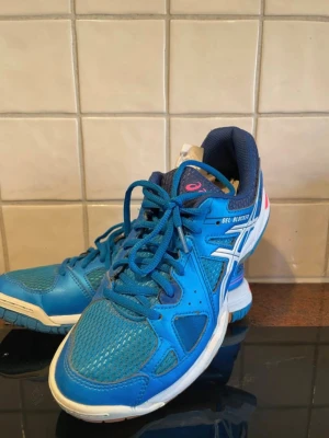Blå Asics inomhusskor - Säljer ett par blå Asics inomhusskor med vita och mörkblå detaljer. Skorna har snörning, ventilerande meshpartier och slitstark sula som passar perfekt för sporter inomhus. Snygg design med Asics-logga på sidan och bekväm vadderad insida.