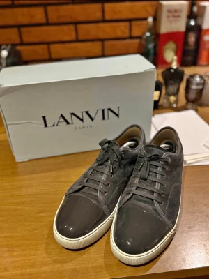 Svarta Lanvin sneakers i mocka - Stilrena svarta sneakers från Lanvin med ovandel i mocka och tåparti i blankt läder. Skorna har vita gummisulor och svarta skosnören. Klassisk låg modell som passar till många olika outfits. Levereras med originalkartong och extra skosnören.
