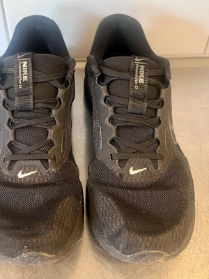 Svarta Nike sneakers  - Snygga svarta Nike sneakers med meshmaterial och svarta syntetdetaljer. Skorna har vita små swoosh-loggor på tån och en större på sidan. Klassisk snörning och låg profil gör dem perfekta för en sportig look.