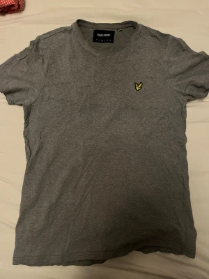Grå t-shirt från Lyle & Scott - Säljer en enkel och stilren grå t-shirt från Lyle & Scott med deras klassiska gula logga på bröstet. T-shirten har rund hals och korta ärmar, perfekt till vardags. Tillverkad i mjuk bomull för skön känsla.