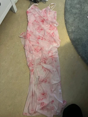 Lång rosa blommig klänning - Säljer en lång, tunn rosa klänning med blommigt mönster i rött. Klänningen har smala axelband, transparent material och volangdetaljer längs hela plagget. Perfekt för sommarens alla tillfällen.