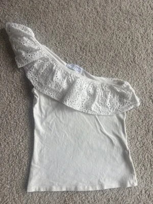 Vit offshoulder topp med volang - Säljer en vit offshoulder topp med bred volang i spetsdetalj. Toppen är ärmlös och har en asymmetrisk design med en axel bar. Perfekt för dig som vill ha en söt och trendig look till sommaren.Skriv om du vill köpa så sätter jag på köp nu.
