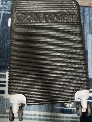 Svart resväska från Calvin Klein - Snygg svart hård resväska från Calvin Klein med räfflad yta och vita detaljer. Väskan har fyra hjul, TSA-lås och dragkedja. Perfekt storlek för weekendresor och smidig att rulla med sig. Stort Calvin Klein-tryck på framsidan.