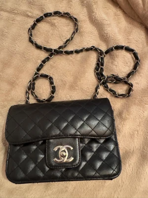 Svart quiltad axelväska  - Säljer en svart quiltad axelväska  med kedjeband i silver och läder. Väskan har röd insida med dragkedjefack. Perfekt för dig som vill ha en tidlös och elegant accessoar.