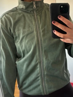 Grön ziptröja från Palm Angels - Grön ziptröja från Palm Angels med hög krage och hel dragkedja framtill. Tröjan har en sportig look, diskret logga på bröstet och är gjord i ett mjukt syntetmaterial. Perfekt för dig som gillar streetwear och enkel stil.