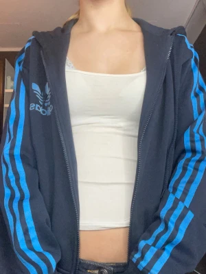 Oversized mörkblå Adidas hoodie med blå ränder - Säljer en mörkblå Adidas hoodie med dragkedja och huva. Klassiska blå ränder längs ärmarna och stor Adidas-logga på bröstet. Hoodien har en avslappnad passform och är gjord i mjukt material, perfekt för en sportig och chill stil.