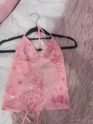 Rosa glittrig  topp - Supersöt rosa topp med blommigt mönster och glittriga paljettdetaljer. Toppen har öppen rygg med snörning och knyt i nacken. Perfekt för dig som vill sticka ut med en feminin och trendig look.