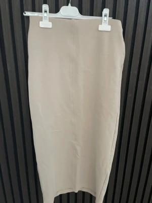 Beige långkjol i mjukt material - Stilren beige långkjol med rak passform och enkel design. Kjolen är tillverkad i ett mjukt och stretchigt material som ger en bekväm känsla. Perfekt att matcha med både sneakers och boots för en trendig look. Aldrig använd 