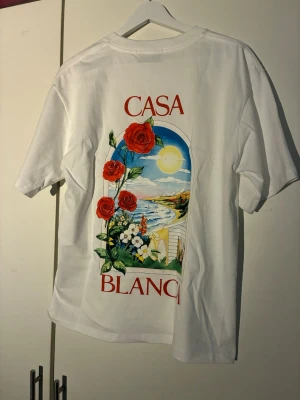Vit Casa Blanca t-shirt med tryck - Vit t-shirt med färgglatt tryck på ryggen som visar röda rosor, hav och solnedgång. Texten 'CASA BLANCA' i rött. Klassisk passform och korta ärmar, perfekt för en avslappnad stil.