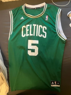 Grön Celtics basketlinne Adidas - Säljer en grön basketlinne från Adidas med Celtics-tryck och nummer 5 på framsidan. På baksidan står det Garnett och samma nummer. Linnet har vita detaljer vid ärmhål och hals, samt NBA-logga på bröstet. Perfekt för basketfans!