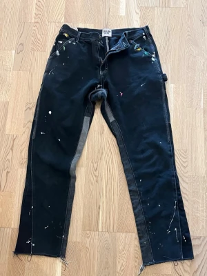 Svarta jeans från Gallery Dept. - Unika svarta jeans från Gallery Dept. med färgstänk och råa kanter nertill. Jeansen har kontrastsömmar och detaljer i grått längs insidan av benen. Perfekta för dig som gillar en kreativ och edgy stil.