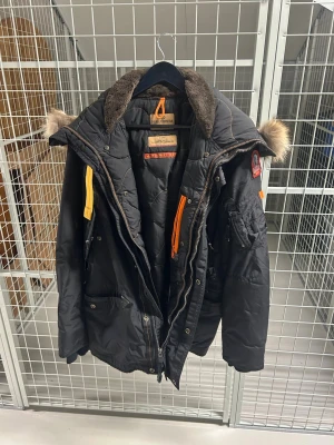 Svart dunjacka från Parajumpers - Säljer en svart dunjacka från Parajumpers med pälsdetalj på huvan, orange detaljer och patch på ärmen. Jackan har flera fickor, dragkedja och tryckknappar. Perfekt för kalla vinterdagar och riktigt snygg streetstil.
