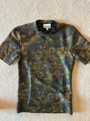Glittrig camo t-shirt från Ganni - Säljer en glittrig t-shirt från Ganni med camouflage-mönster i gröna, blå och guldiga toner. T-shirten har korta ärmar och en klassisk rund halsringning. Perfekt för dig som vill sticka ut lite extra.