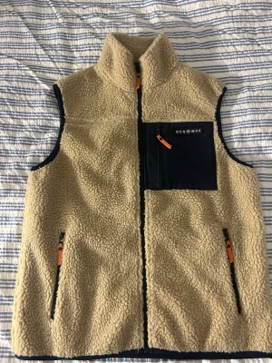 Beige fleeceväst med fickor OCNWKS - Mysig beige fleeceväst från OCNWKS med hög krage och dragkedja. Västen har två sidofickor med svarta dragkedjor och orange detaljer samt en svart bröstficka med dragkedja. Perfekt för lager-på-lager och chill stil. Kommer ej till använding för mig då den är för stor.