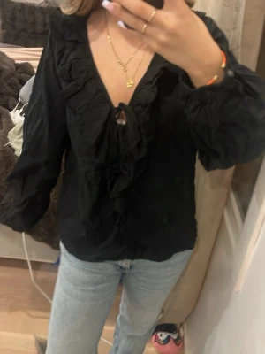 Svart blus med volang och knyt - Snygg svart blus med djup v-ringning, volangkant och knytband framtill. Långa puffiga ärmar och lätt skrynkligt tyg ger en avslappnad men chic look. Perfekt att styla med jeans eller kjol för en trendig outfit.