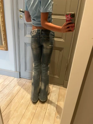 Blå bootcut jeans med slitningar - Säljer ett par ljusblå bootcut jeans med så slitna detaljer och klassisk femficksmodell. Jeansen har en låg midja och en patch på bakfickan. Perfekta för en avslappnad och trendig look.