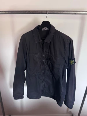 Svart overshirt från Stone Island - Stilren svart overshirt från Stone Island med dragkedja framtill och klassisk krage. Jackan har två bröstfickor och den ikoniska Stone Island-loggan på vänster ärm. Perfekt för lager-på-lager och streetwear-stil.
