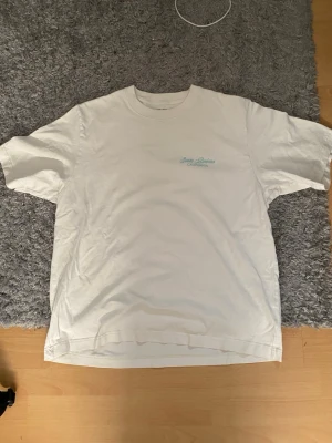 Vit t-shirt med Santa Barbara-tryck - Vit t-shirt med turkost tryck på bröstet där det står 'Santa Barbara California'. Klassisk passform och rund halsringning. Perfekt för en avslappnad stil och enkel att matcha med jeans eller shorts.