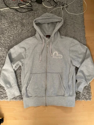 Grå zip hoodie från Palace och evisu - Snygg grå zip hoodie från Palace med broderad logga på bröstet. Hoodien har dragsko i huvan, två stora fickor framtill och ribbade muddar vid ärmar och midja. Perfekt för en chill och avslappnad stil. Tillverkad i mjukt bomullsmaterial.