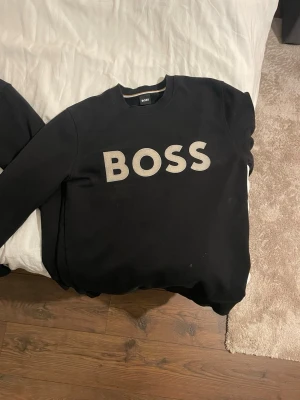Svart sweatshirt från BOSS - Svart sweatshirt från BOSS med stor vit logga över bröstet. Klassisk rund halsringning och ribbade muddar vid ärmslut och nederkant. Perfekt för en avslappnad och stilren look.