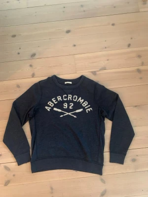 Mörkblå sweatshirt Abercrombie & Fitch - Mörkblå sweatshirt från Abercrombie & Fitch med vit text och siffran 92 samt två korsade åror på bröstet. Tröjan har rund halsringning och ribbade muddar vid ärmslut och nederkant. Den här dessutom lite små slitningar runtom halsen.