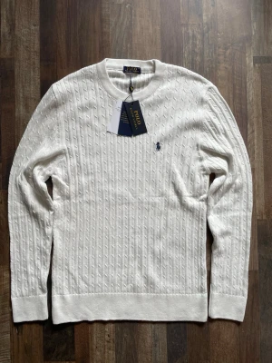 Vit kabelstickad tröja - Kabelstickad vit tröja med rund halsringning och diskret broderad logga på bröstet. Tröjan har långa ärmar och ribbade muddar vid ärmslut och nederkant. Klassisk och stilren design i mjukt material.