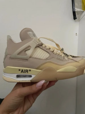 Off-White x Air Jordan 4 Sail sneakers - Snygga Off-White x Air Jordan 4 Sail sneakers i beige och krämvitt. Skorna har meshpaneler, detaljer i plast och texten 'AIR' på sulan. Klassiska Off-White detaljer som zip-tie och text på skosnören. Perfekt för dig som gillar streetwear och unika samarbeten.