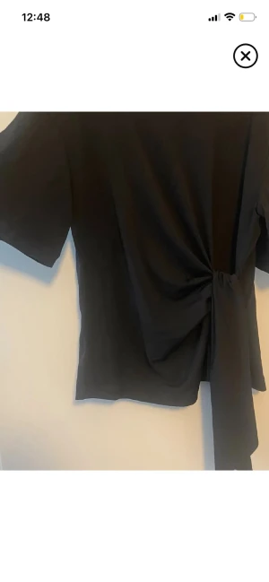 Svart topp med knyt i midjan - Snygg svart topp med korta ärmar och trendig knytning i midjan. T-shirten har en enkel, stilren design och är tillverkad i mjukt bomullstyg. Perfekt för dig som gillar minimalistisk stil med en twist. Andvänd en gång så o fint skick