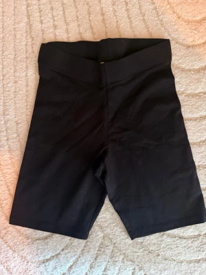 Svarta cykelshorts med hög midja - Säljer ett par svarta cykelshorts med hög midja. De är stretchiga och sitter tight, perfekta till sommaren eller träning. Materialet känns mjukt och följsamt, och längden går till strax ovanför knäna. Enkla att matcha med olika toppar.