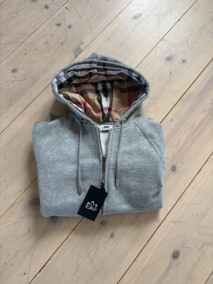 Grå hoodie med dragkedja och mönstrad huva - Säljer en helt oanvänd burberry zip hoodie då den var förliten, 600kr pris ej huggit i sten. Ny pris 1700.