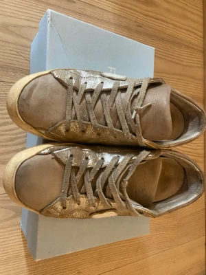 Guldglittriga sneakers Philippe Model - Snygga sneakers från Philippe Model i guldglittrigt material med beige detaljer och vita inslag på sidan. Skorna har klassisk låg profil, rund tå och snörning framtill. Perfekta för dig som vill sticka ut med lite extra bling i vardagen.
