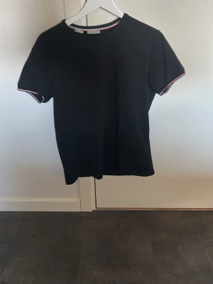 Svart Moncler t-shirt med rödvit kant - Svart t-shirt från Moncler med korta ärmar och rund hals. Ärmsluten har snygga rödvita ränder och ett Moncler-märke sitter på ena ärmen. Tillverkad i mjuk bomull för skön känsla och clean look.