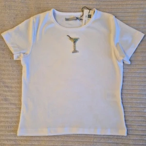 Beaded Vit t-shirt med pärlad martiniglasdetalj - Snygg vit t-shirt med korta ärmar och en unik pärlad martiniglasdetalj på bröstet. Perfekt för dig som vill ha något extra i garderoben. Tillverkad i mjuk bomull för skön känsla hela dagen.