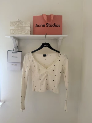 Vit ribbad topp - Söt vit ribbad topp med små svarta hjärtan, v-ringad och croppad modell. Toppen har lång ärm och knappar framtill. Perfekt för dig som gillar gulliga detaljer och en trendig look.