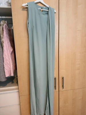 Ljusblå långklänning med drag över axeln - Elegant ljusblå långklänning i ett slätt, mjukt material. Klänningen är ärmlös med rund halsringning och har ett långt, draperat släp framtill som ger en stilren och modern look. Perfekt för speciella tillfällen när du vill sticka ut.