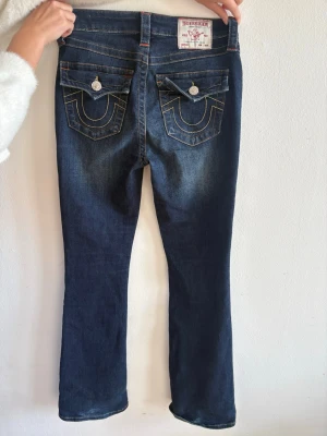 Mörkblå bootcut jeans True Religion - Snygga mörkblå bootcut jeans från True Religion med kontrastsömmar och ikoniska fickdetaljer bak. Jeansen har fem fickor, knappgylf och coola vita knappar på bakfickorna. Materialet är klassisk denim med lätt stretch för skön passform.