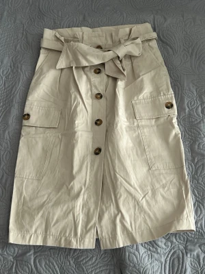 Snygg beige kjol med knappar och bälte H&M - Stilren beige kjol från H&M med knappar framtill, stora fickor på sidorna och ett matchande bälte i midjan. Kjolen är tillverkad i bomull och har en rak passform som ger en snygg och avslappnad look.