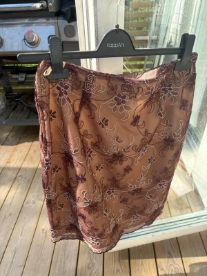 Blommig tvådelad spetsklänning i brunt/vinrött och beige - Sött tvådelat set med kjol och topp i transparent vinröd spets med blommiga broderier över beige tyg. Toppen har korta ärmar och v-ringning med dekorativ rosett. Kjolen är rak och följsam. Perfekt för dig som gillar romantisk och feminin stil.