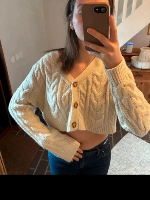 Cropped kabelstickad kofta i offwhite - Trendig cropped kofta i offwhite med kabelstickat mönster och stora träknappar framtill. Koftan har v-ringning, långa ärmar och ribbade muddar. Perfekt för lager på lager och ger en cool vibe till din outfit.