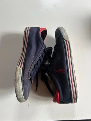 Ralph Lauren sneakers - Snygga marinblå sneakers med röda detaljer och broderad logga på sidan. Skorna har vita och röda ränder längs sulan och svarta skosnören. Ovandelen är i tyg och insidan har en röd kontrast. Perfekta för en avslappnad streetstil.