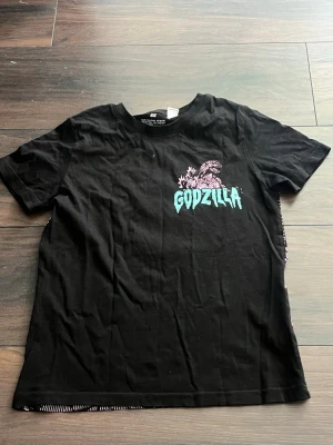 Svart Godzilla t-shirt från H&M - Säljer en svart t-shirt från H&M med ett coolt Godzilla-tryck i lila och turkos på bröstet. T-shirten har rund hals och korta ärmar, perfekt för dig som gillar streetwear och popkultur.