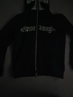 Svart zip hoodie med tryck - Säljer en svart hoodie med dragkedja och huva. På bröstet står det 'Cross Angel' i gotisk stil och på ryggen finns ett stort kors i silverglittrande material. Hoodien har långa ärmar och ribbade muddar. Perfekt för dig som gillar streetwear och edgy stil.