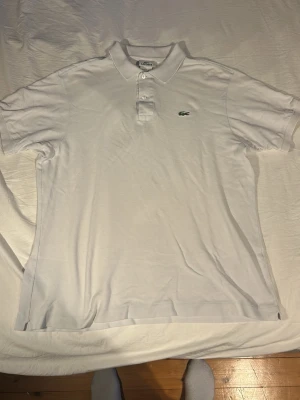 Vit pikétröja från Lacoste - Klassisk vit pikétröja från Lacoste med korta ärmar och krage. Tröjan har knappar framtill och den ikoniska gröna krokodilloggan på bröstet. Tillverkad i bomull för en skön och luftig känsla.