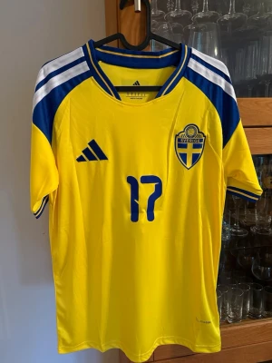 Sverige fotbollströja nr 17 Gyökeres - Gul och blå officiell Sverige fotbollströja från Adidas med nummer 17 och namnet Gyökeres på ryggen. Tröjan har korta ärmar, blå detaljer på axlar och krage samt det svenska landslagets emblem på bröstet. Perfekt för match eller träning.