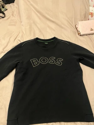 Svart sweatshirt från BOSS - Svart sweatshirt från BOSS med stor logga i Guld på bröstet. Tröjan har rund halsringning, långa ärmar och diskreta sömdetaljer vid axlarna. Perfekt för en clean och stilren look.
